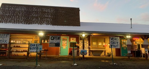 A rustic storefront with signs for various goods, illuminated at dusk, featuring an "OPEN" sign and snow on the roof.