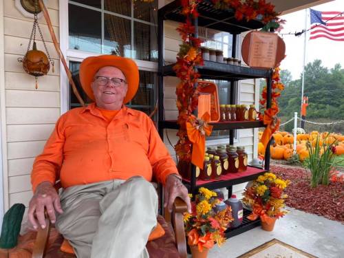 A man in an orange hat and shirt sits on a porch surrounded by fall decorations and jars, with pumpkins in the background.
