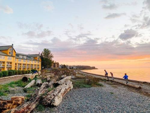 Children play along a pebbly beach at sunset, with a yellow building and logs scattered along the shore.