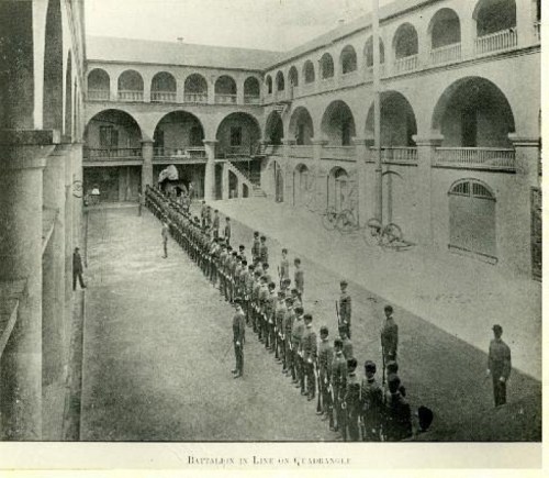 Historic image of a battalion lined up in a courtyard, surrounded by arched architecture and large doors.