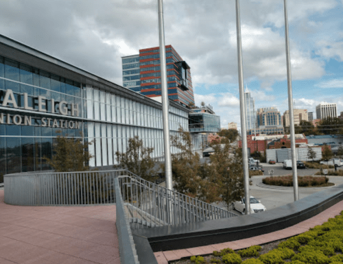 View of Raleigh Union Station with modern buildings and flags in the foreground, under a cloudy sky.