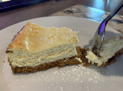 A slice of creamy cheesecake on a plate, with a fork partially cutting into it, and a dusting of powdered sugar on top.