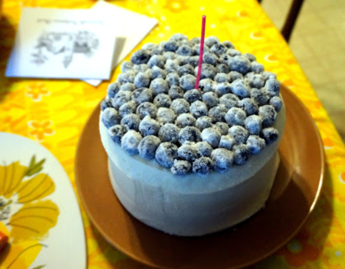 A round cake topped with blueberries and a single candle, placed on a brown plate with a yellow tablecloth in the background.