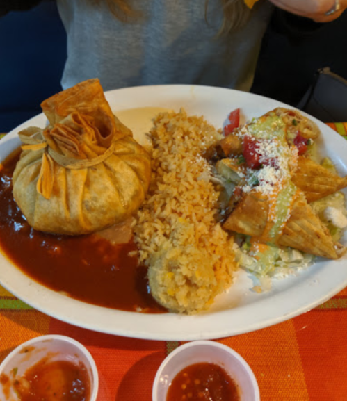 A plate of Mexican food featuring a stuffed pastry, rice, and crispy tacos with toppings, accompanied by dipping sauces.