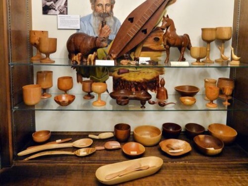 A display of wooden crafts, including bowls, spoons, and carved figures, with a portrait of a man in the background.