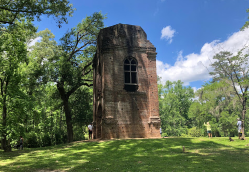 A tall, brick tower stands amidst lush green trees under a blue sky with scattered clouds. People are visible nearby.