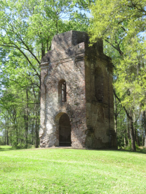 A weathered brick structure stands among lush green trees, surrounded by grass in a serene outdoor setting.