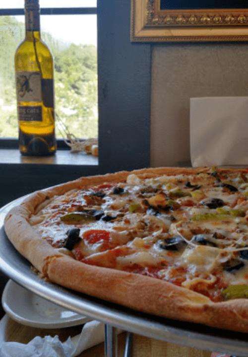 A close-up of a pizza topped with vegetables, cheese, and olives, with a bottle of wine in the background.