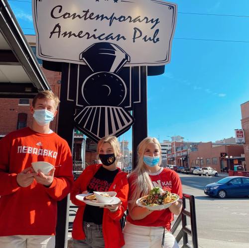Three people in masks hold plates of food in front of a pub sign, smiling on a sunny day.