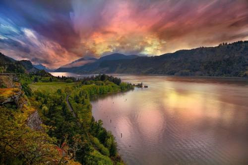 A vibrant sunset over a river, surrounded by lush greenery and mountains, with dramatic clouds in the sky.