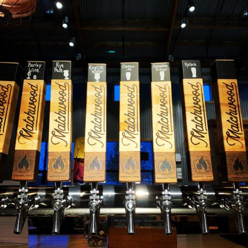 A row of wooden beer taps with labels for various brews at a brewery, set against a rustic backdrop.
