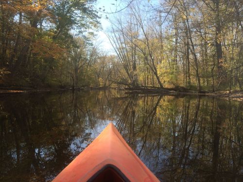 A kayak's bow on a calm river, surrounded by autumn trees reflecting in the water under a clear blue sky.