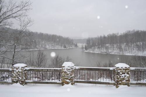 Snow-covered landscape with a river and trees in the background, viewed from a stone railing. Snowflakes are falling.
