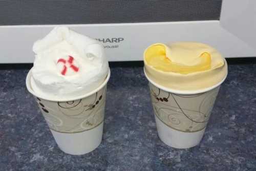 Two paper cups with whipped cream: one topped with red sprinkles and the other with a yellow swirl.