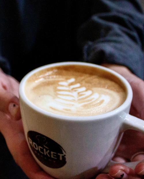 A person holding a white coffee cup with latte art on top, featuring a leaf design.