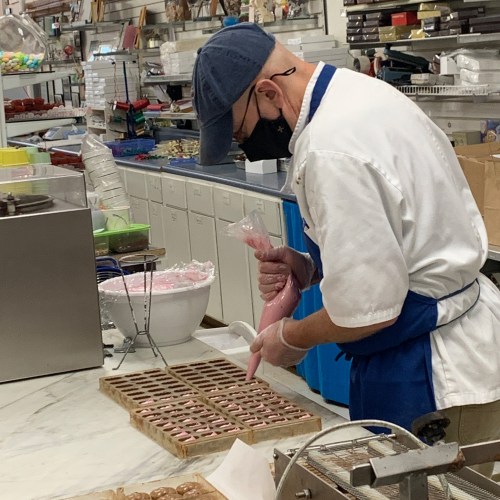 A person in a kitchen decorates pastries with pink icing, surrounded by baking supplies and trays of treats.