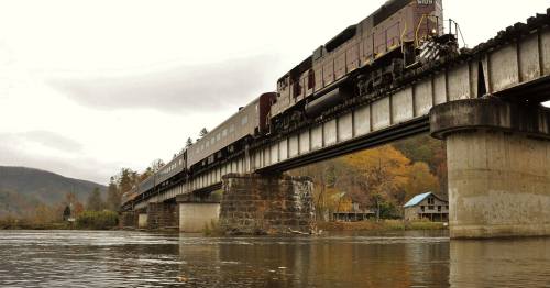A train crosses a bridge over a river, surrounded by autumn trees and a distant building.