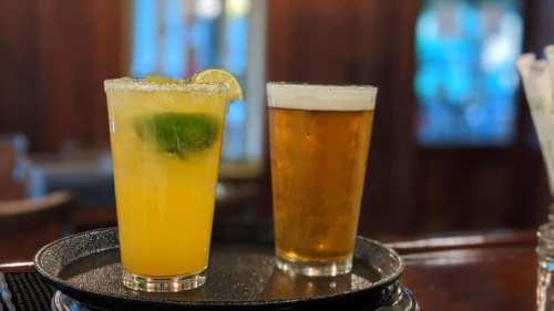 A tray holds a margarita with lime and a pint of beer, set against a blurred bar background.