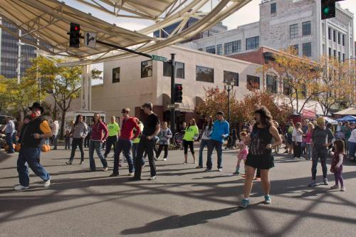 A lively street scene with people dancing and enjoying a community event under a canopy, surrounded by buildings and trees.