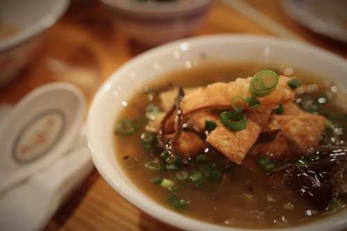 A bowl of soup with crispy toppings, green onions, and mushrooms, served on a wooden table.