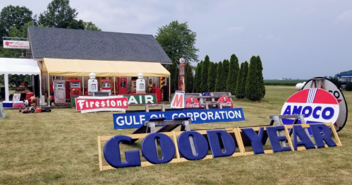 A vintage gas station display featuring old signs for Firestone, Gulf Oil, Amoco, and Goodyear on a grassy field.