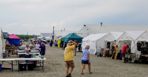 A bustling outdoor market with people browsing tents and tables filled with various goods under a cloudy sky.