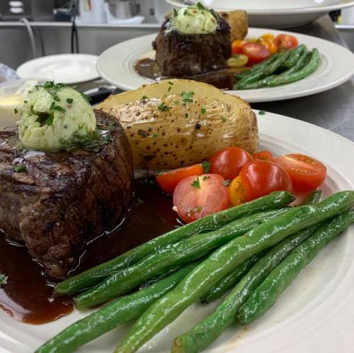 A plated meal featuring a steak with herb butter, green beans, roasted potatoes, and cherry tomatoes, garnished with parsley.