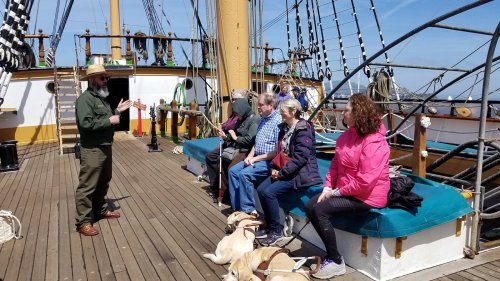 A park ranger speaks to a group of people seated on a ship's deck, with dogs resting nearby under a clear blue sky.