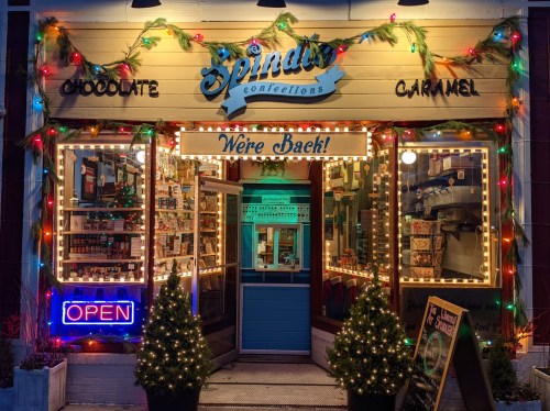 A festive candy shop entrance adorned with lights and decorations, featuring a sign that says "We're Back!" and an "Open" neon sign.