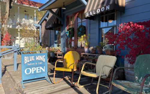 A charming blue building with a saloon and café sign, featuring colorful chairs and potted plants on a wooden deck.