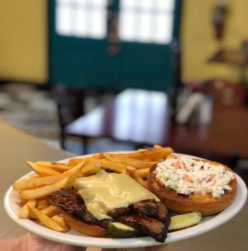 A plate with grilled steak topped with cheese, fries, a bun with coleslaw, and a pickle slice on the side.