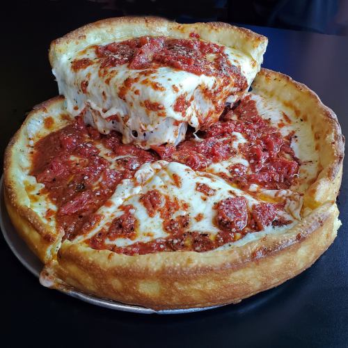A deep-dish pizza with a thick crust, topped with melted cheese and marinara sauce, served on a black table.