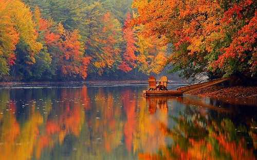 A serene lake surrounded by vibrant autumn foliage, with two chairs on a wooden dock reflecting in the water.