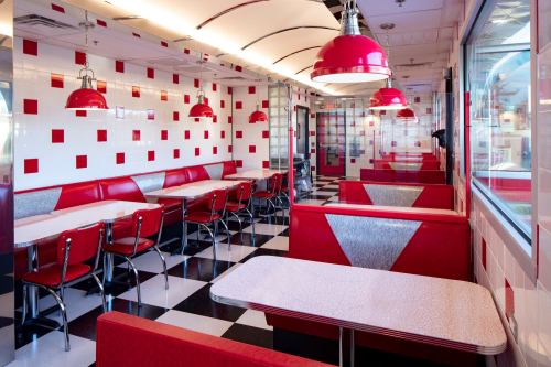 A retro diner interior featuring red and white decor, checkered floors, and red pendant lights.