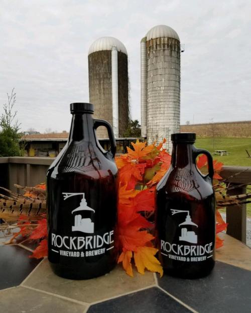 Two dark glass growlers with "Rockbridge Vineyard & Brewery" logo, surrounded by autumn leaves and silos in the background.