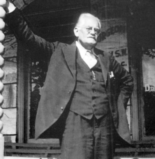 A historical black-and-white photo of a man in a suit, standing with one arm raised, in front of a building.