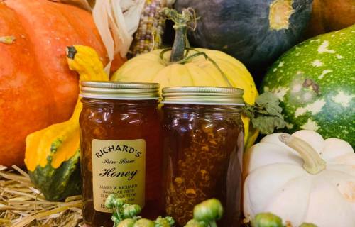 Two jars of honey sit in front of colorful pumpkins and gourds, surrounded by straw.