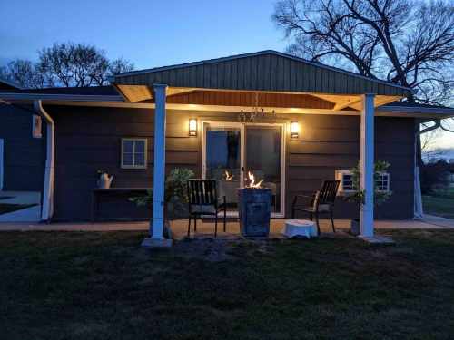 A cozy house at dusk with a welcoming porch, two chairs, and a fire pit, surrounded by trees.