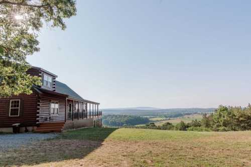 A log cabin with a porch overlooks a vast green valley and distant mountains under a clear blue sky.