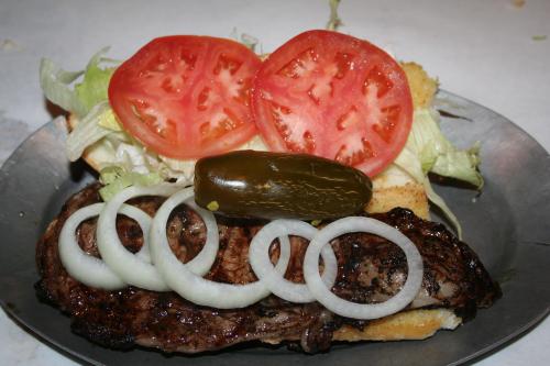 A close-up of a sandwich with grilled meat, lettuce, tomato slices, onion rings, and a pickle on a metal plate.
