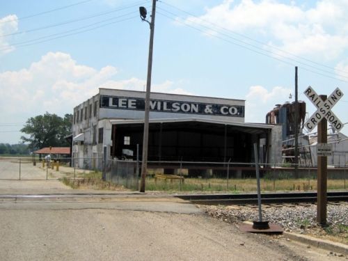 Abandoned building with "Lee Wilson & Co." sign, near a railroad crossing and open field under a blue sky.