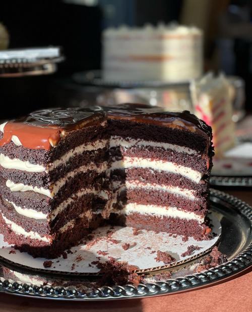 A chocolate layer cake with creamy filling, partially sliced, on a silver platter with another cake in the background.