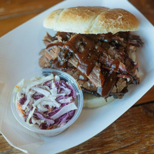 A pulled pork sandwich with barbecue sauce on a bun, served with a side of coleslaw in a small container.