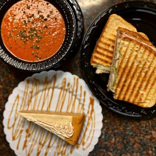 A bowl of tomato soup, a grilled sandwich, and a slice of cake on a decorative plate.