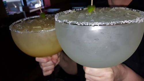 Two large margarita glasses filled with ice, one with a salted rim and a lime garnish, held by a person.