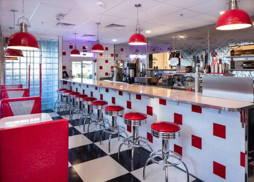 A retro diner interior featuring red stools, checkered floor, and bright red pendant lights.