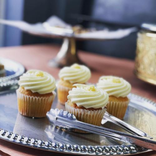 A silver tray holds three cupcakes with cream frosting, accompanied by a pair of serving tongs.