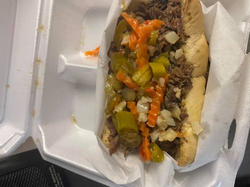 A sandwich filled with shredded beef, topped with pickled peppers, onions, and jalapeños, served in a takeout container.