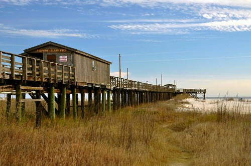 A wooden pier extends over sandy beach, leading to a small building under a clear blue sky. Grassy dunes frame the scene.
