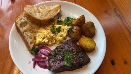 A plate with scrambled eggs, grilled steak, roasted potatoes, pickled onions, and two slices of toasted bread.